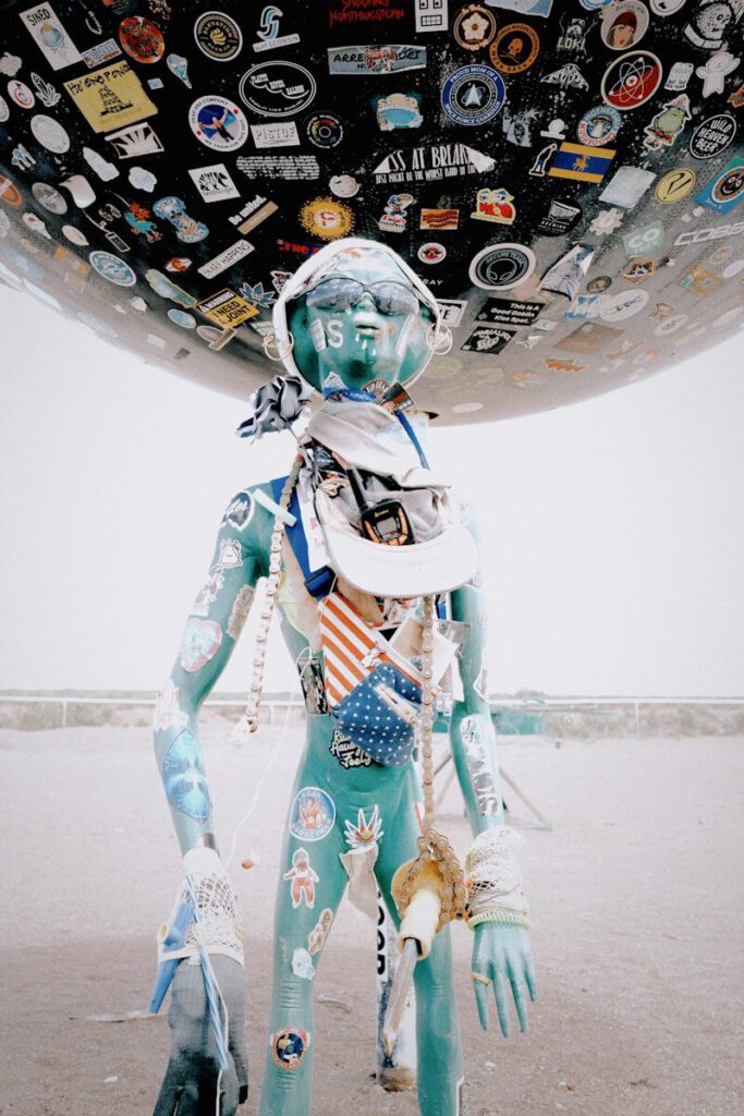 A turquoise mannequin with stickers under a large sticker-covered sphere.