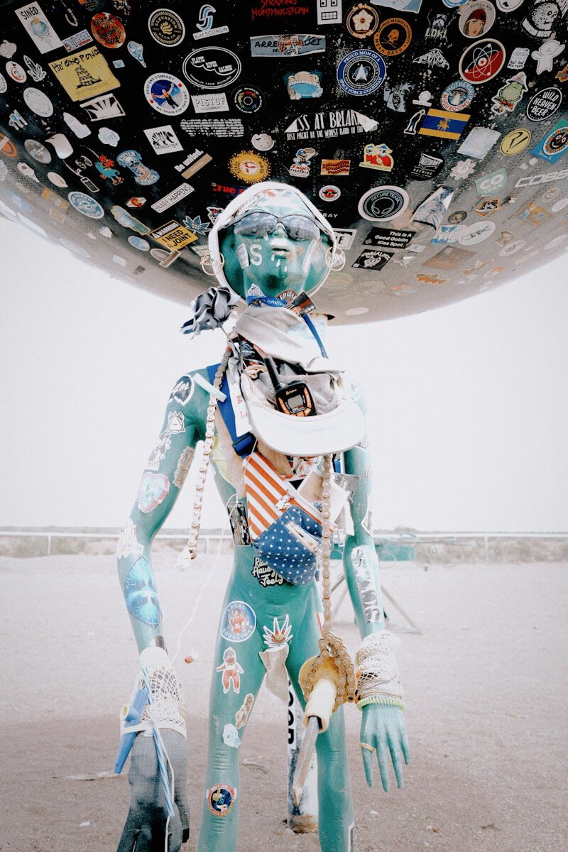 A turquoise mannequin with stickers under a large sticker-covered sphere.